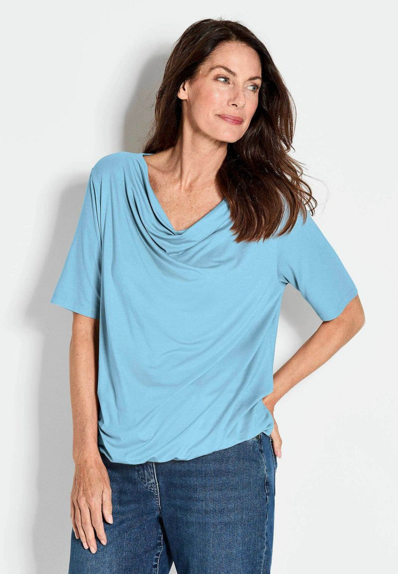 Lichtblauw kort mouwtop met een gedrapeerde halslijn, gemaakt van zachte stof. Het shirt heeft een relaxte pasvorm en een subtiele textuur. Gecombineerd met denim jeans.