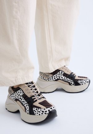 Zapatillas gruesas con plataforma con patrones moteados y a rayas en beige, negro y blanco, combinadas con pantalones color crema sobre un fondo claro.