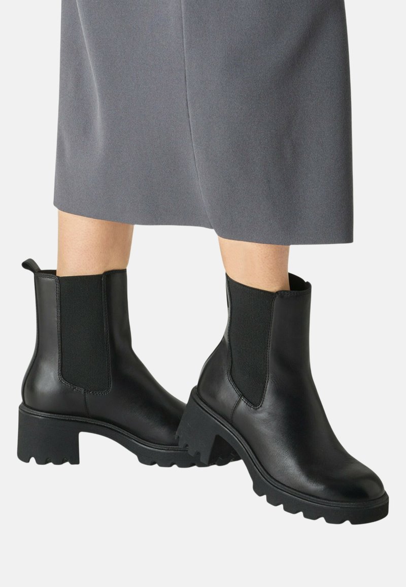 Femme portant des bottines en cuir noir à talons épais, avec des panneaux latéraux élastiques et une jupe grise mi-longue.