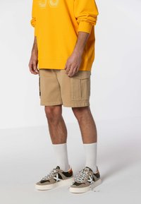 Felpa gialla, pantaloni cargo beige con tasche laterali, calzini bianchi e scarpe da ginnastica verde/grigio con accenti e lacci bianchi.