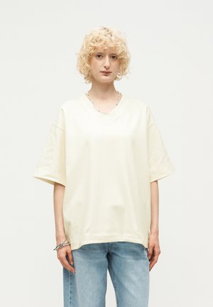 Άτομο με κοντά σγουρά ξανθά μαλλιά που φοράει μια υπερμεγέθη κοντομάνικη πουκάμισο εκρού και ανοιχτό μπλε τζιν, με φόντο απλό.