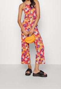Mono floral con patrones multicolores en rojo, amarillo y morado; diseño de pierna ancha. Bolso amarillo y sandalias de plataforma negras.