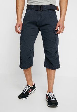 Mand iført mørkeblå cargoshorts med sort bælte og sorte sneakers med hvide snørebånd og røde detaljer, stående på en lys gulv.