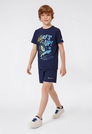 Niño caminando, vistiendo una camiseta gráfica azul marino con el texto "Surf's Up!", pantalones cortos azul marino y zapatillas blancas con cordones azules sobre un fondo claro y sencillo.