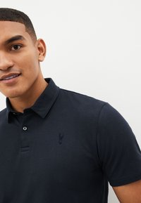 Marineblaues Polohemd aus weichem Stoff mit umgeschlagenem Kragen, das ein dezentes gesticktes Logo auf der Brust und eine Knopfleiste aufweist.