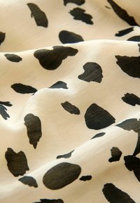 Světlé béžové látky s černým vzorem kravského tisku, který obsahuje různé nepravidelné tvary a velikosti. Textura vypadá hladce a lehce leskle.