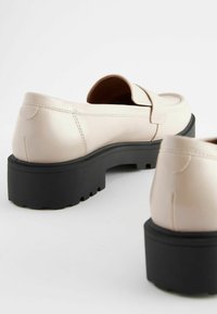 Beige nahkaiset liukuhäntäsandaalit, joissa on pyöristetty kärki, musta paksu pohja ja koristeellinen hihna. Lihava pinta, minimalismi muotoilu.