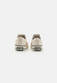 Bež Converse All Star tenisice s niskim dizajnom, kožnim i platnenim materijalom, crnim naglaskom i gumiranim potplatima. Stražnji pogled prikazuje detalje logoa.