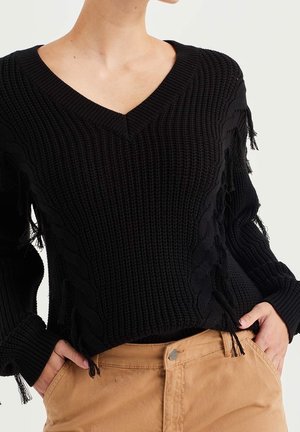 Persoon draagt een zwarte, grofgebreide V-hals trui met kwastjes, gecombineerd met een beige broek met hoge taille, handen in de zakken, tegen een effen achtergrond.