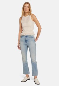 Débardeur en tricot beige avec une texture côtelée, associé à un jean évasé bleu clair avec des ourlets effilochés, et des chaussures blanches à enfiler.