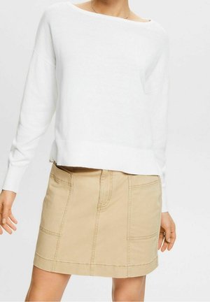 Kobieta ubrana w biały sweter z długim rękawem i beżową spódnicę typu A z przednimi kieszeniami, stojąca na tle jednolitego jasnego tła.