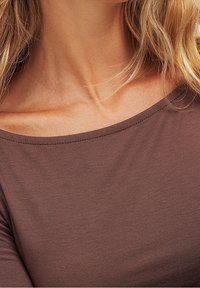 Ruskea, istuva toppi, jossa on leveä, pyöreä pääntie ja sileä pinta; näyttää osan kaulasta ja olkapäästä, ei näkyviä varusteita tai kuvioita.