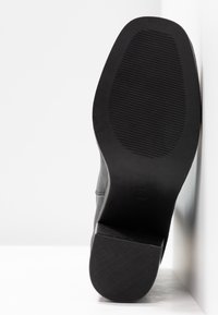 Semelle de chaussure en cuir noir avec grip texturé et petit talon rectangulaire, photographiée par le dessous sur fond blanc.