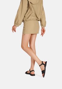 Giacca a vento beige con cordino, abbinata a una gonna corta. Sandali neri con cinturini intrecciati e chiusura a fibbia sul retro. Design con suola piatta.