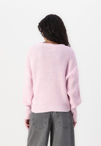 Růžový pletený svetr s volným střihem, širokými rukávy a žebrovaným lemem. Nošený s šedými volnými kalhotami, které mají texturovaný vzor.