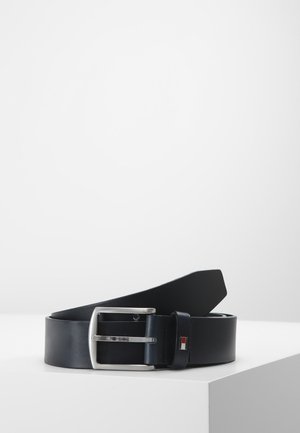 Schwarzer Ledergürtel mit einer silbernen, rechteckigen Schnalle und einem kleinen rechteckigen Logo in Rot, Weiß und Blau an der Gürtelschlaufe, präsentiert auf einer weißen Oberfläche.