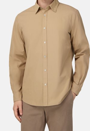 Muž nosící béžovou košili s dlouhým rukávem s límcem a hnědé kalhoty, stojící s jednou rukou v kapse.