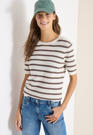 Ung kvinde smilende, iført en grøn kasket, hvid og brunstribet kortærmet sweater og blå jeans med den ene hånd i lommen.