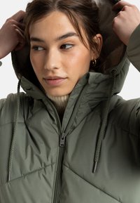 Veste matelassée verte avec capuche, présentant un design matelassé, une fermeture éclair et des cordons de serrage. La doublure intérieure est de couleur claire, ajoutant un contraste.