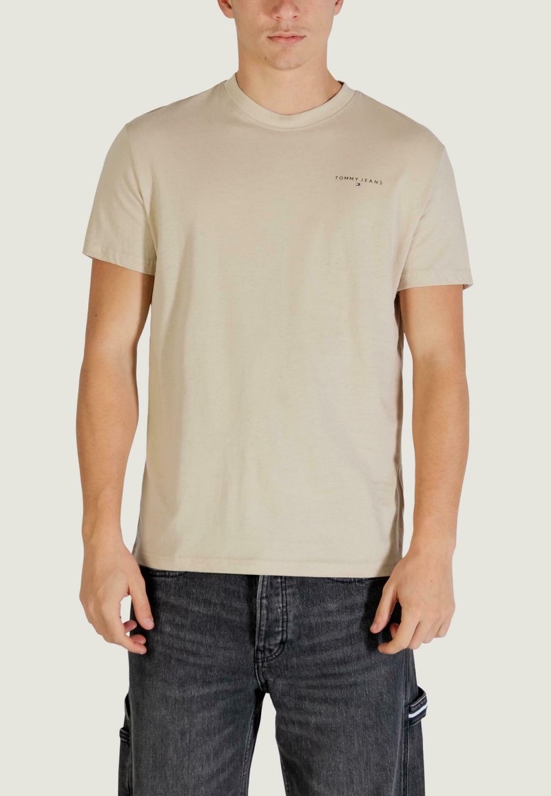 Beiges Baumwoll-T-Shirt mit Rundhalsausschnitt, kurzen Ärmeln und kleinem Logo auf der Brust. Kombiniert mit dunkelblauen Jeans.