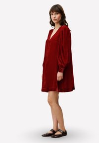 Rode fluwelen jurk met een diepe V-hals, lange opgeblazen mouwen en een losse, A-lijn silhouet. Model draagt donkere platte schoenen.