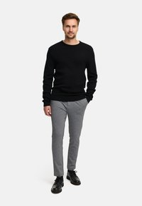 Czarny żebrowany sweter, szare dopasowane spodnie i czarne sznurowane buty. Model stoi na tle w jednolitym kolorze.