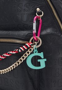 Černá taška z umělé kůže s texturou, zdobená zlatým zipem, růžovým karabinou, splétaným přívěskem na klíče v červené, černé a bílé barvě a tyrkysovým přívěskem ve tvaru písmena "G".
