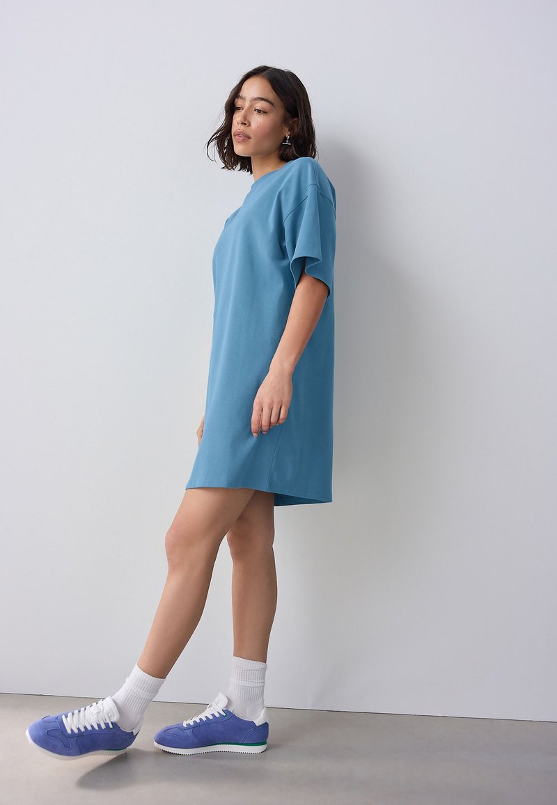 Rochie tip tricou supradimensionată de culoare albastru deschis, realizată dintr-un material moale, cu mâneci scurte și croială lejeră, asortată cu adidași mov și șosete albe.