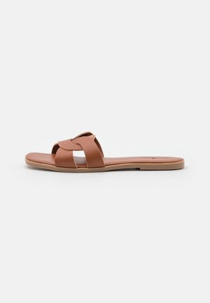 Brun flad slip-sandal med brede, snoede læderstropper og en tynd sål på en enkel lys baggrund.