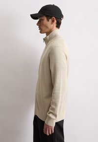 Beige zip-up trui met een gestructureerde brei, hoge kraag en geribde manchetten, gecombineerd met een zwarte pet; model staat in profiel tegen een lichte achtergrond.