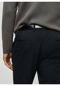 Schwarze Hose mit glatter Textur, ausgestattet mit Gesäßtaschen und einem eleganten schwarzen Gürtel. Kombiniert mit einem grauen Langarmshirt.