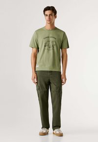 Camiseta de algodón verde oliva con estampado gráfico y cuello redondo, combinada con pantalones cargo oliva que tienen bolsillos laterales y un corte relajado.