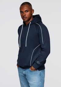 Mężczyzna w granatowej bluzie z kapturem z białymi lamówkami i jasnoniebieskich dżinsach, stojący z rękami w kieszeniach na jednolitym tle.
