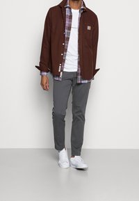 Brązowa kurtka zapinana na guziki nałożona na koszulę w kratę, biała koszulka, szare spodnie i białe trampki. Cechy to kieszenie oraz kontrastowe wzory.