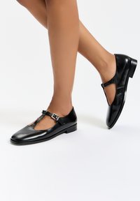 Musta nahkainen Mary Jane -kenkä, jossa on pyöreä varvas, T-hihna-design ja pieni solkiyksityiskohta. Matala korko ja sileä pinta.