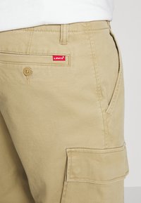 Pantalones cargo beige de tela de mezcla de algodón, con cierre de botón, bolsillo lateral y una etiqueta del logo rojo de Levi's en el bolsillo trasero.