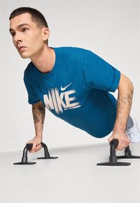 Blå kortærmet t-shirt med hvid "NIKE" grafik. Person der udfører armstræk med sorte armstøtter på en ensfarvet baggrund.