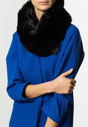 Personne portant un manteau bleu aux manches retroussées et une écharpe en fausse fourrure noire enroulée autour du cou, les bras doucement croisés.