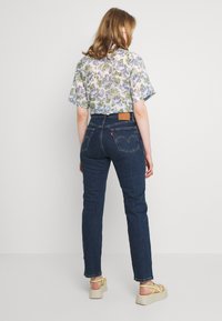 Jeans de mezclilla azul con un diseño ajustado, que cuentan con bolsillos traseros con parche y una etiqueta marrón. Combinados con una blusa corta de manga larga con estampado floral y sandalias.