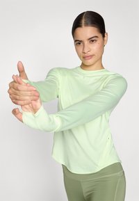 Lys grøn langærmet træningstop med rund hals, glat tekstur, tommelfingerhuller og sidelommer, parret med olivengrønne leggings.