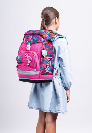 Mädchen mit Pferdeschwanz, das ein hellblaues Kleid und einen pinkfarbenen Rucksack mit Blumenmuster, Flamingo-Patch und reflektierendem Streifen trägt, steht mit dem Rücken zur Kamera.