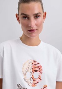 Camiseta blanca de algodón con un gráfico de una calavera en estampado de animal en tonos naranjas y marrones, con texto en un patrón circular alrededor.