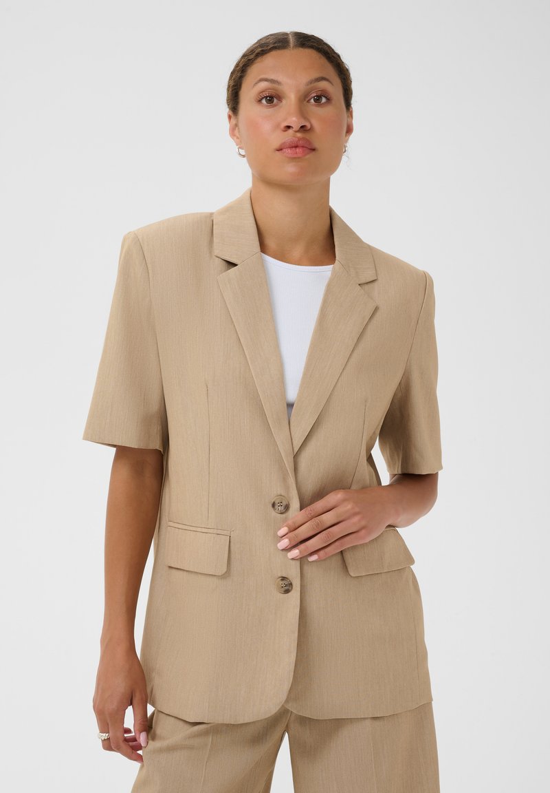 Femme portant un blazer beige à manches courtes sur un haut blanc, debout avec une main reposant sur les boutons du blazer, fond neutre.