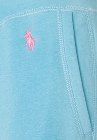 Logo de joueur de polo brodé en rose sur un tissu bleu clair avec des coutures visibles et une ouverture de poche latérale.