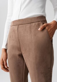 Pantaloni in suede marrone con una texture liscia, taglio su misura e tasche laterali, abbinati a un top bianco aderente a maniche lunghe.