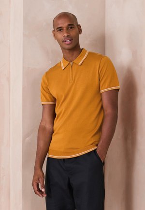 Mann trägt ein senfgelbes kurzärmeliges Poloshirt mit weißem Besatz, steht mit einer Hand in der Tasche vor einer beigen Wand.