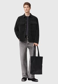 Black corduroy jacket with two front pockets, worn over a gray shirt, paired with light gray jeans and black tote bag. Smooth texture and relaxed fit.