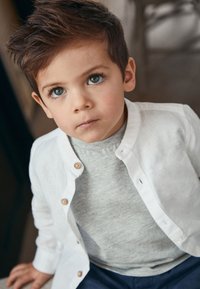 Camicia bianca di lino con bottoni sopra una t-shirt grigio chiaro, pantaloni blu, capelli corti e scuri e grandi occhi azzurri, seduto contro uno sfondo neutro.