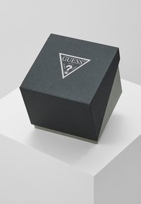 Čierna štruktúrovaná darčeková krabička trojuholníkového tvaru s strieborným logom "GUESS" na vrchu. Predložená na bielom podstavci.