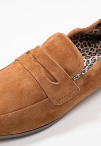 Chaussure slip-on en suede marron avec un col rembourré, un détail de sangle décorative et une doublure en imprimé léopard contrastant. Texture lisse, forme arrondie.